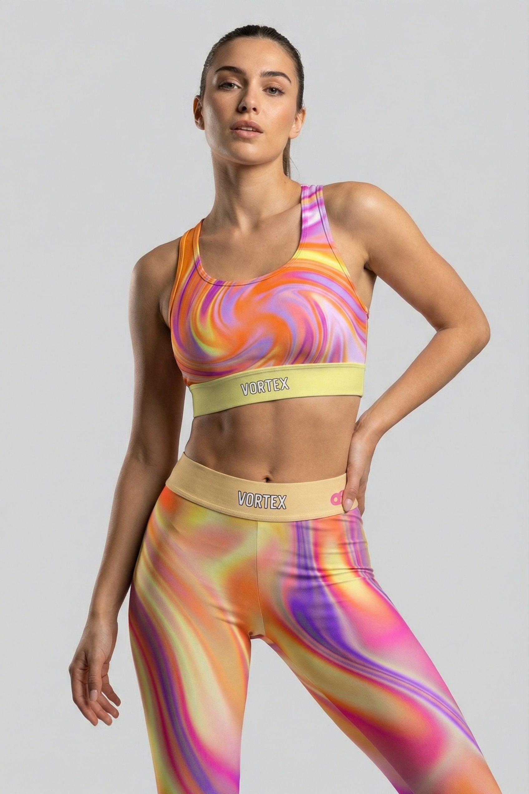 Front view of tie-dye sports bra with orange, purple, and yellow swirl pattern, featuring 'VORTEX' printed on the pastel yellow waistband.