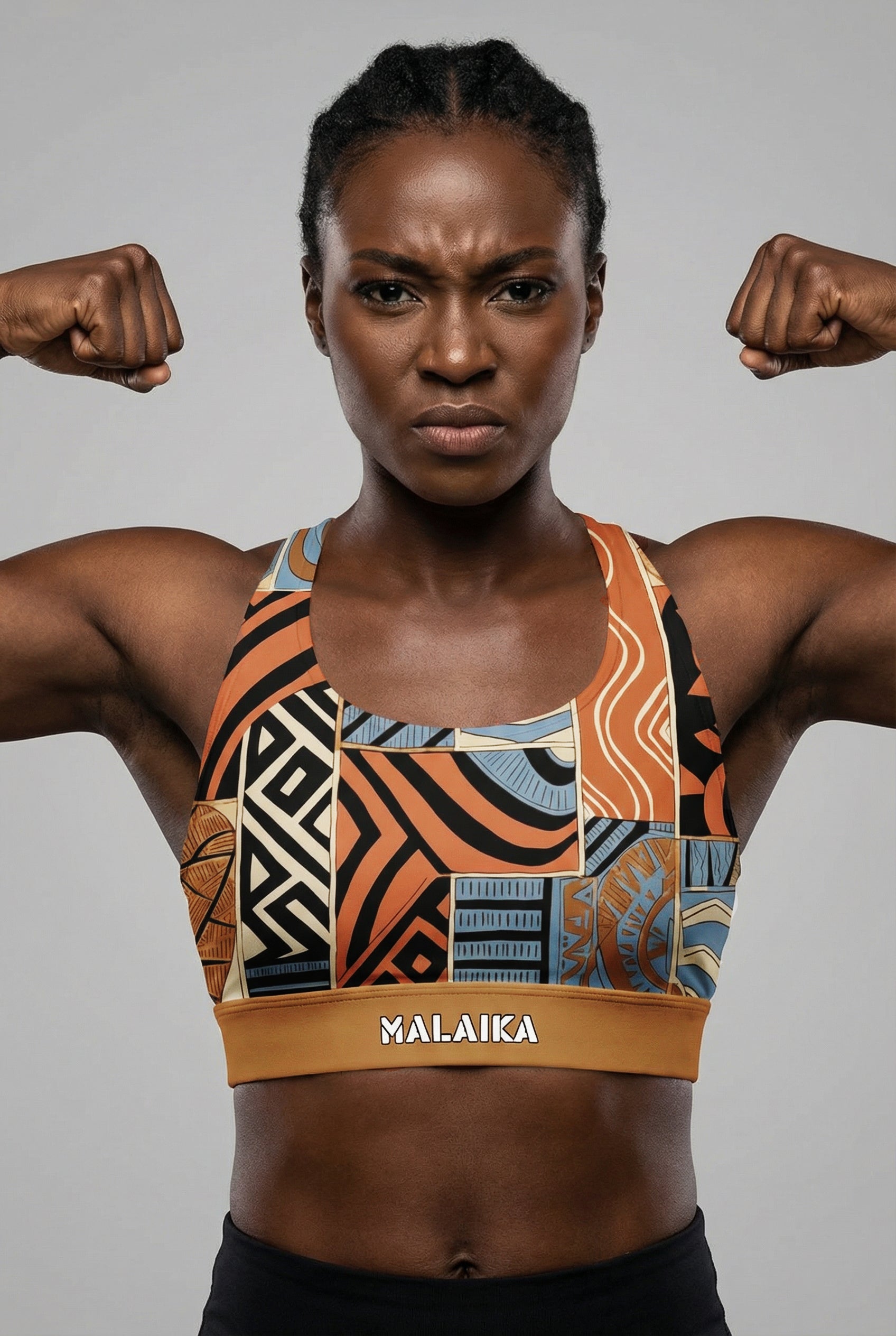 Front view of a fit model flexing biceps, wearing a bold orange and black abstract tribal sports bra with 'MALAIKA' printed on the golden waistband.