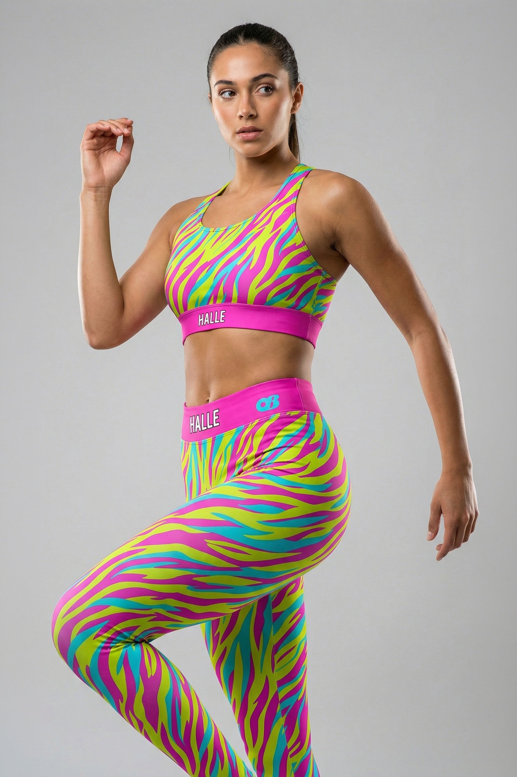 Front view of a confident woman with crossed arms wearing a neon pink, yellow, and cyan tiger stripe sports bra, featuring 'HALLE' printed on the solid hot pink waistband.