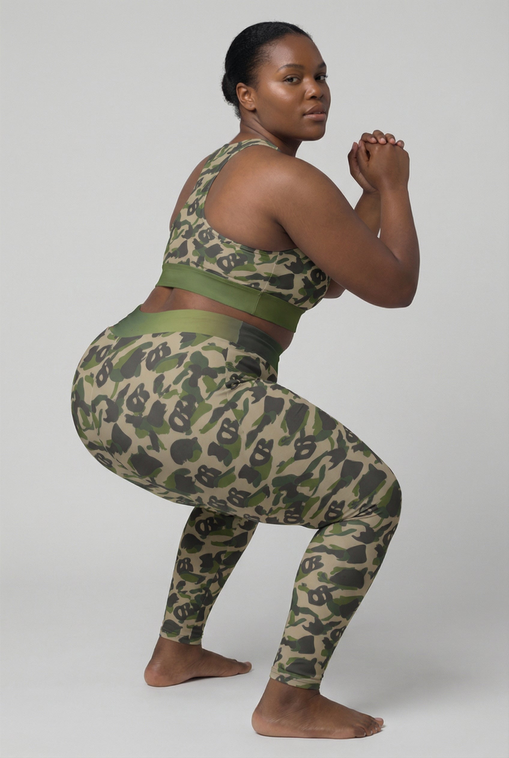Front view of green camouflage sports bra featuring hidden 'OB' monogram shapes in olive and tan, with 'ZIPPY' printed on the olive green waistband.