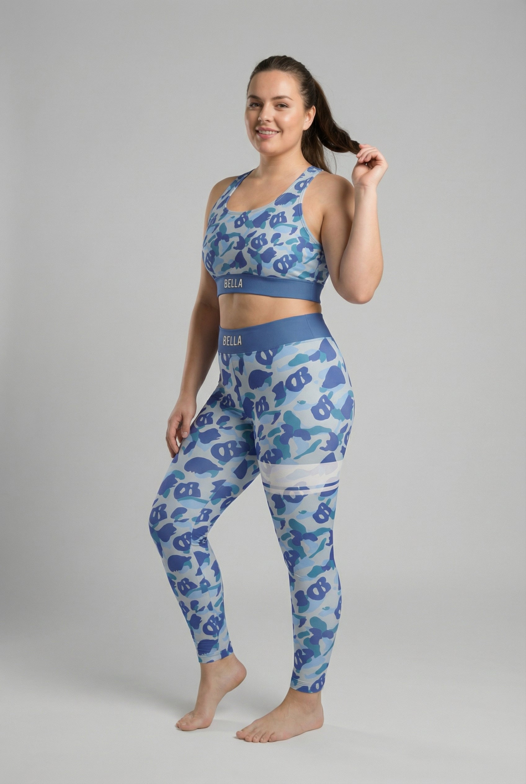 Front view of blue camouflage sports bra featuring hidden 'OB' monogram shapes in sky blue and dark blue, with 'BELLA' printed on the slate blue waistband.