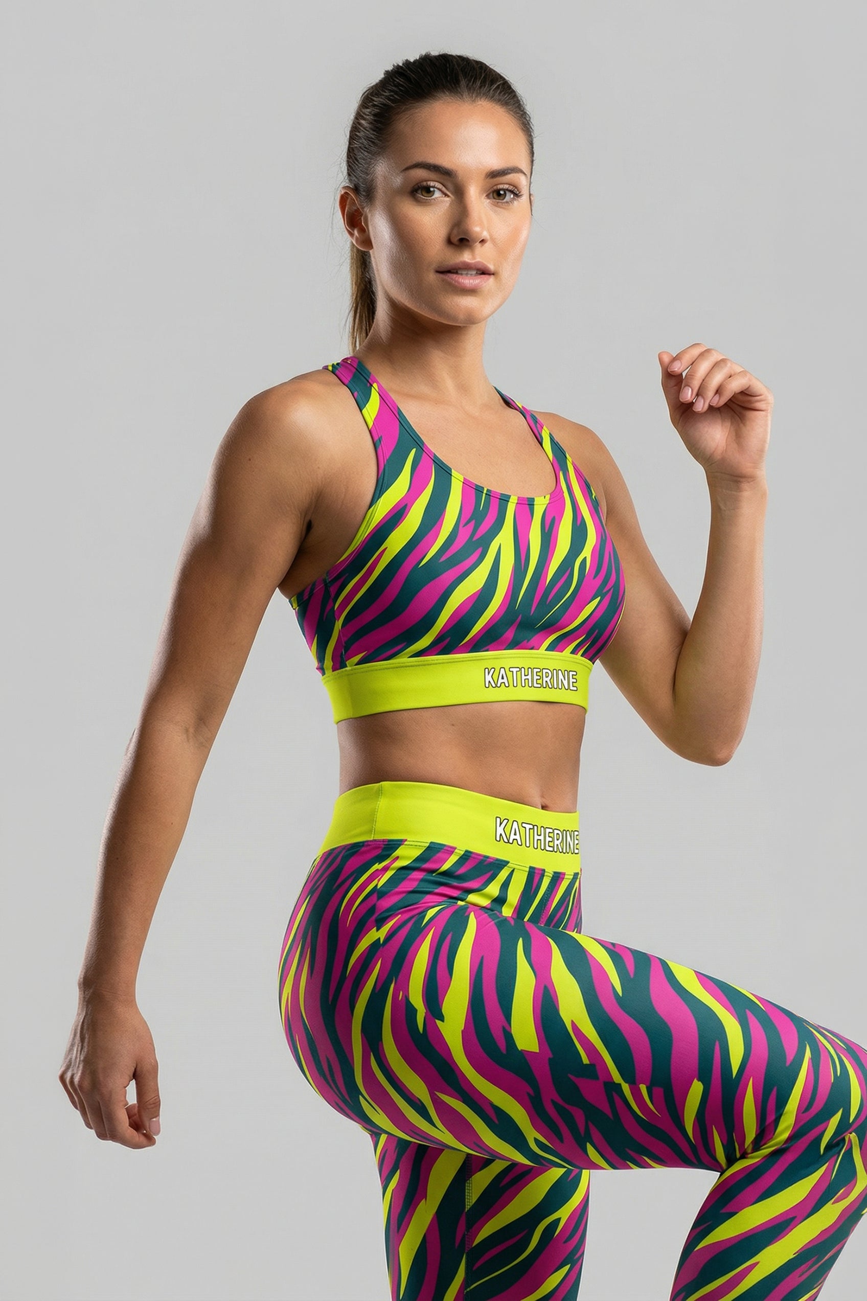 Front view of a confident woman wearing a hot magenta, yellow, and teal tiger stripe sports bra, featuring 'KATHERINE' printed on the solid neon lime waistband.
