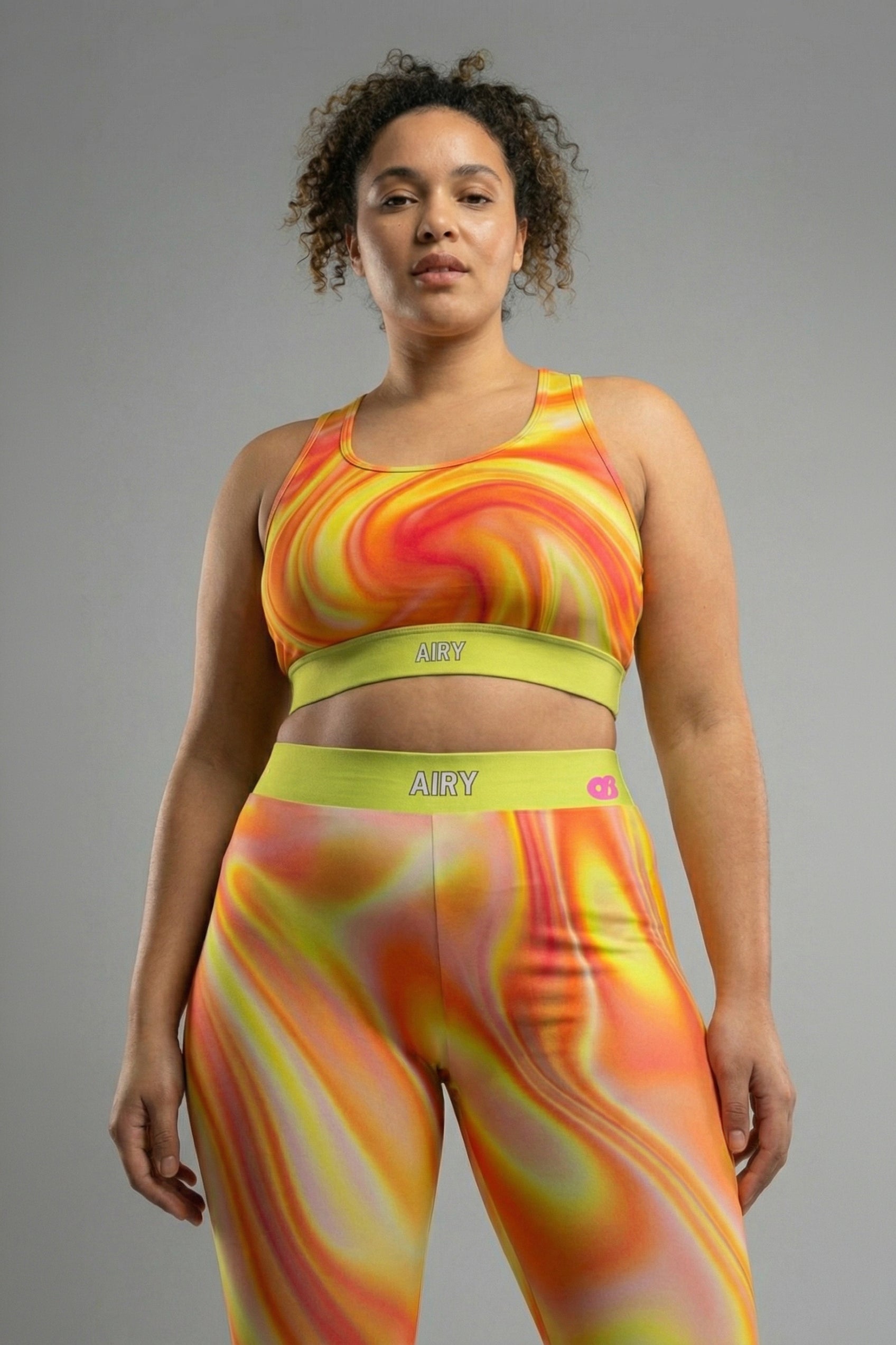 Front view of tie-dye sports bra in orange, yellow, and red swirls, featuring 'AIRY' printed on the neon lime green waistband.