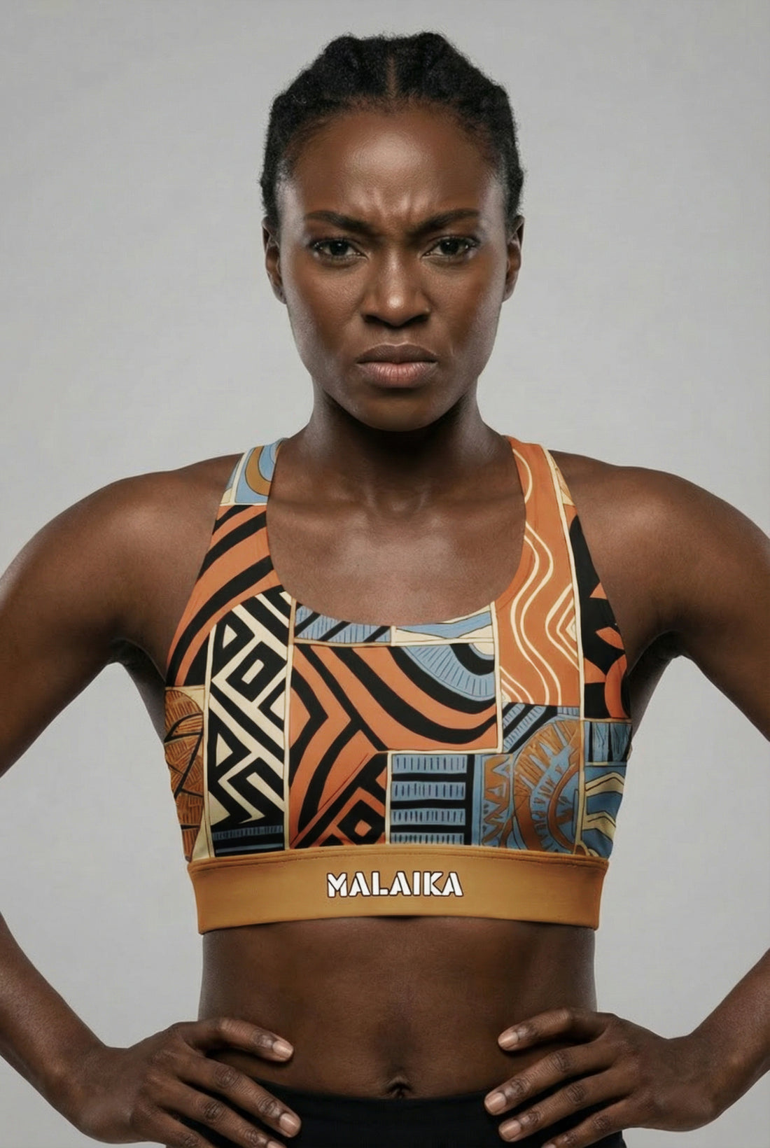 Front view of a fit model flexing biceps, wearing a bold orange and black abstract tribal sports bra with 'MALAIKA' printed on the golden waistband.