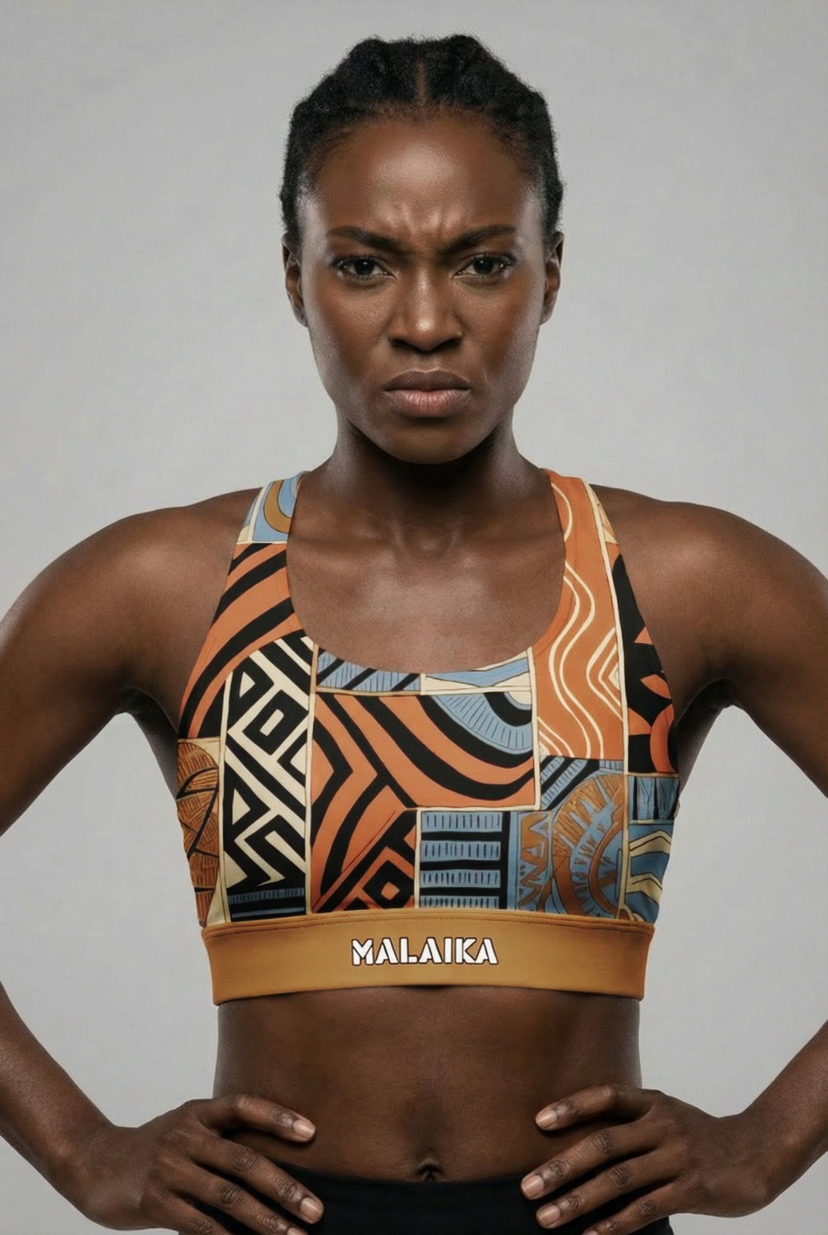 Front view of a fit model flexing biceps, wearing a bold orange and black abstract tribal sports bra with 'MALAIKA' printed on the golden waistband.