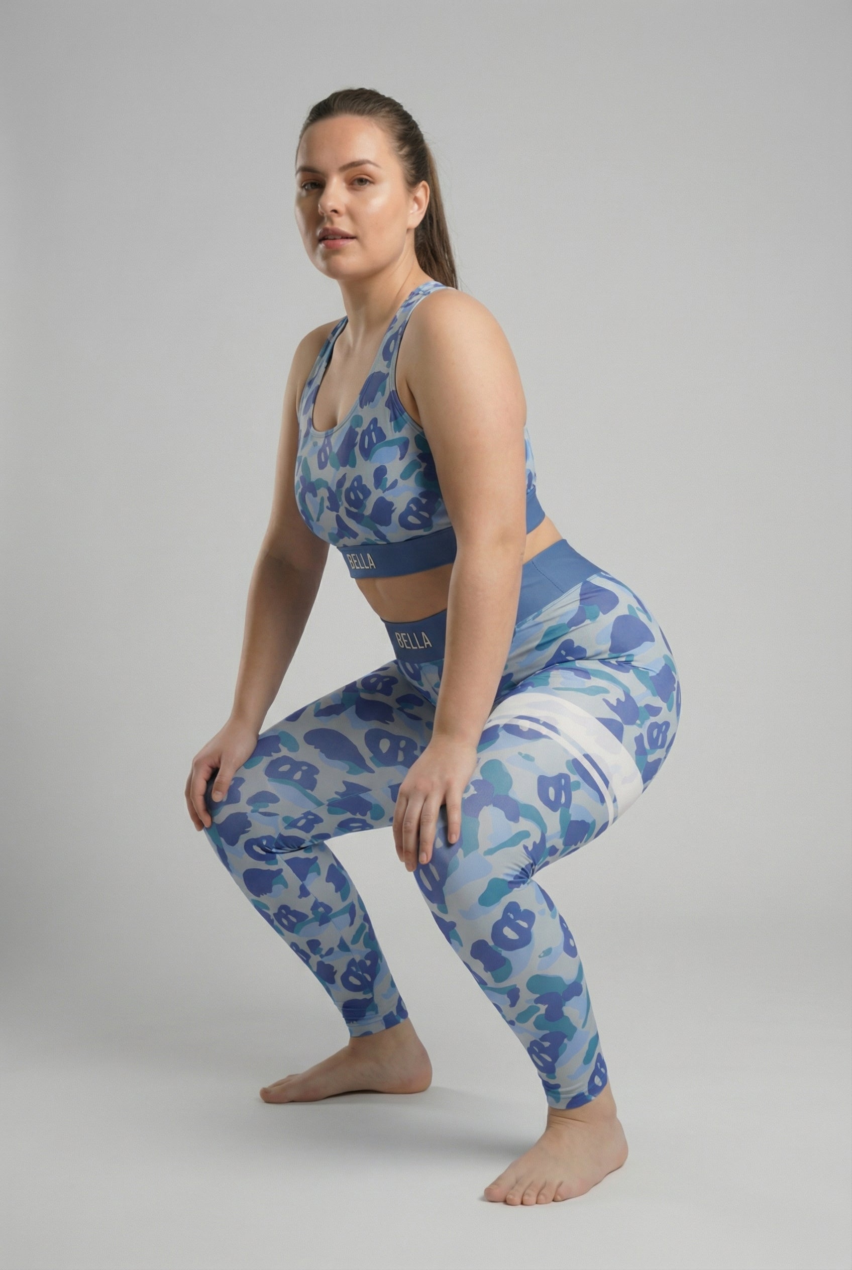 Front view of blue camouflage sports bra featuring hidden 'OB' monogram shapes in sky blue and dark blue, with 'BELLA' printed on the slate blue waistband.