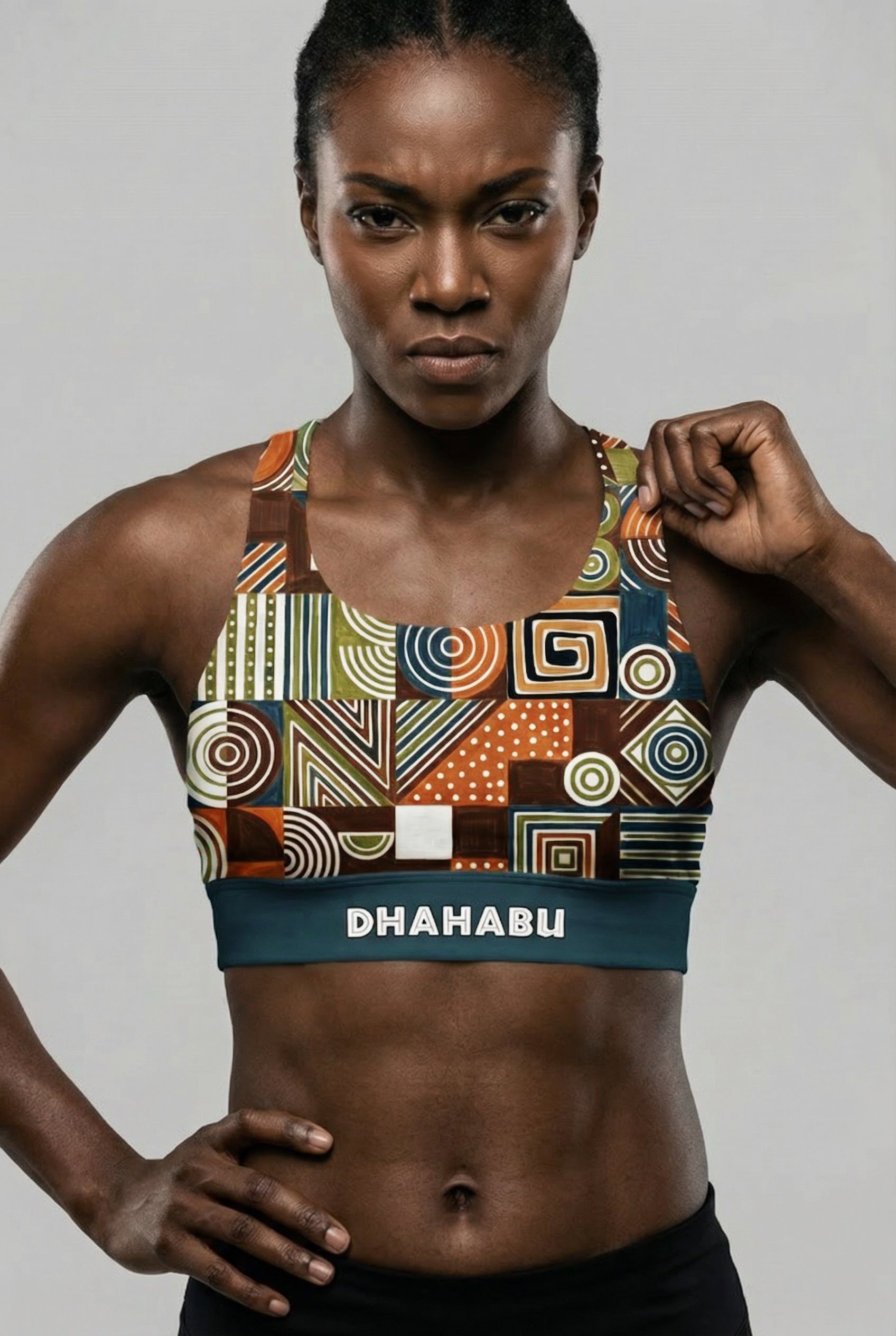 Front view of a dark-skinned model with arms crossed, wearing a tribal geometric pattern sports bra with 'DHAHABU' printed on the teal waistband.