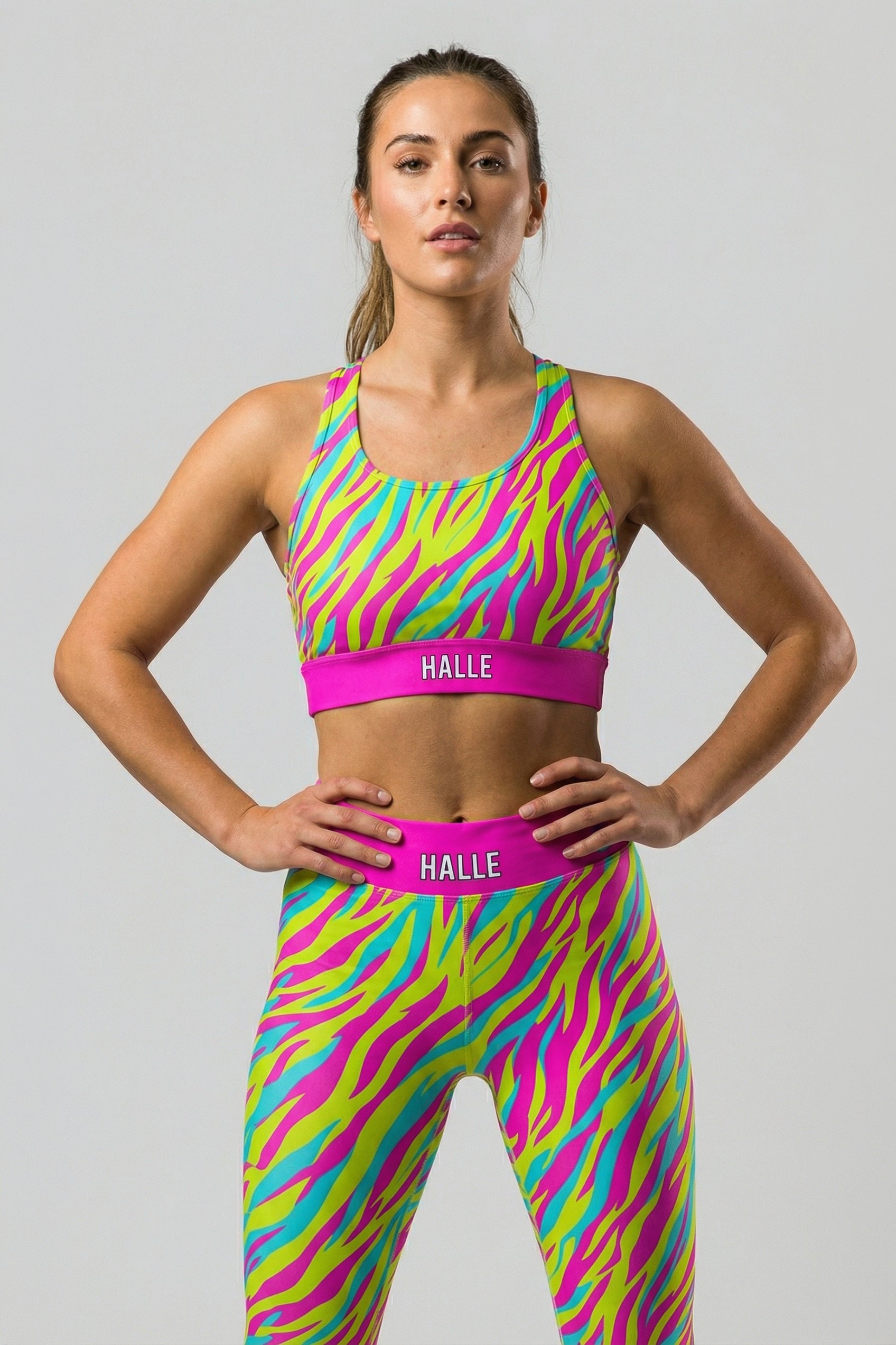 Front view of a confident woman with crossed arms wearing a neon pink, yellow, and cyan tiger stripe sports bra, featuring 'HALLE' printed on the solid hot pink waistband.