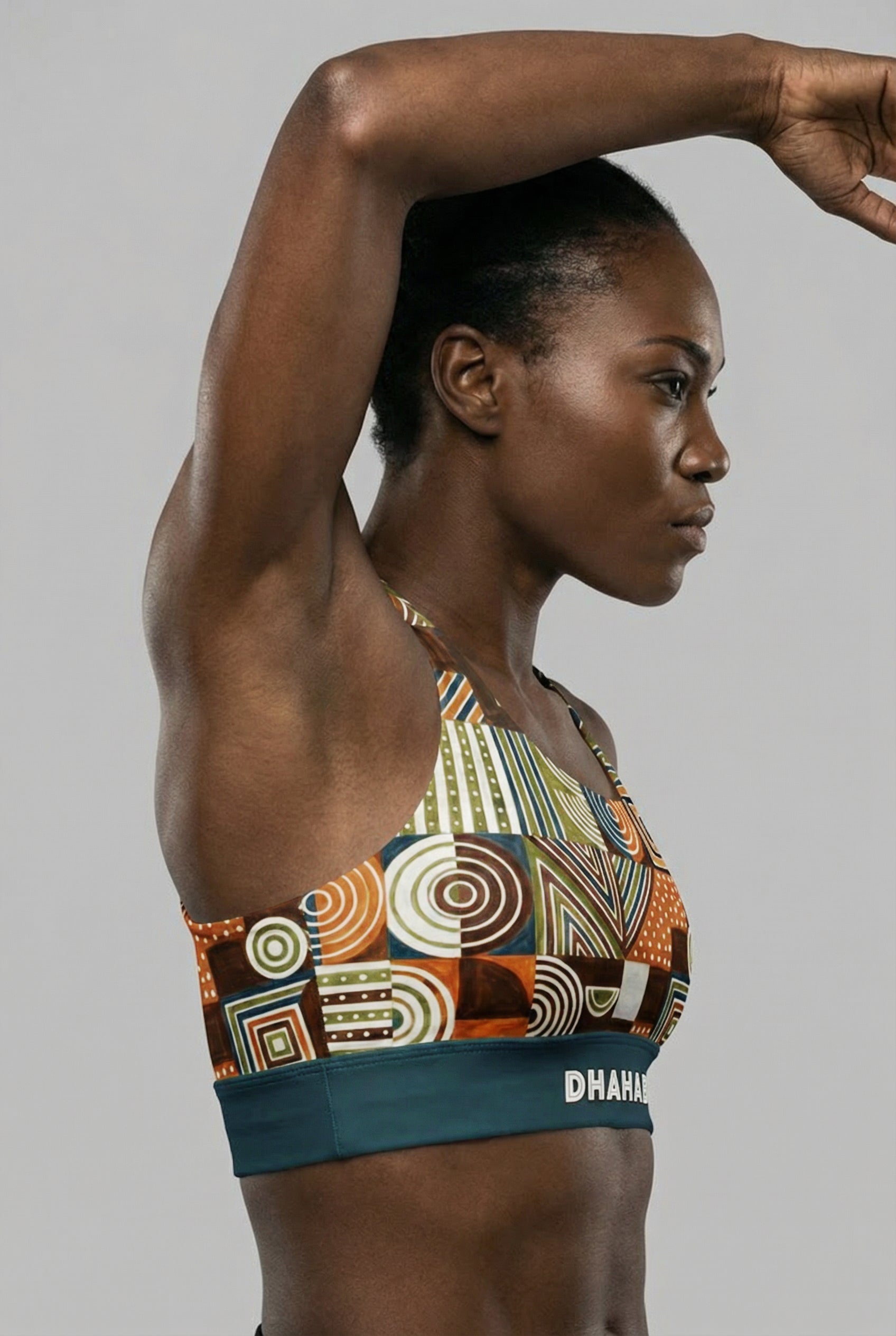 Front view of a dark-skinned model with arms crossed, wearing a tribal geometric pattern sports bra with 'DHAHABU' printed on the teal waistband.