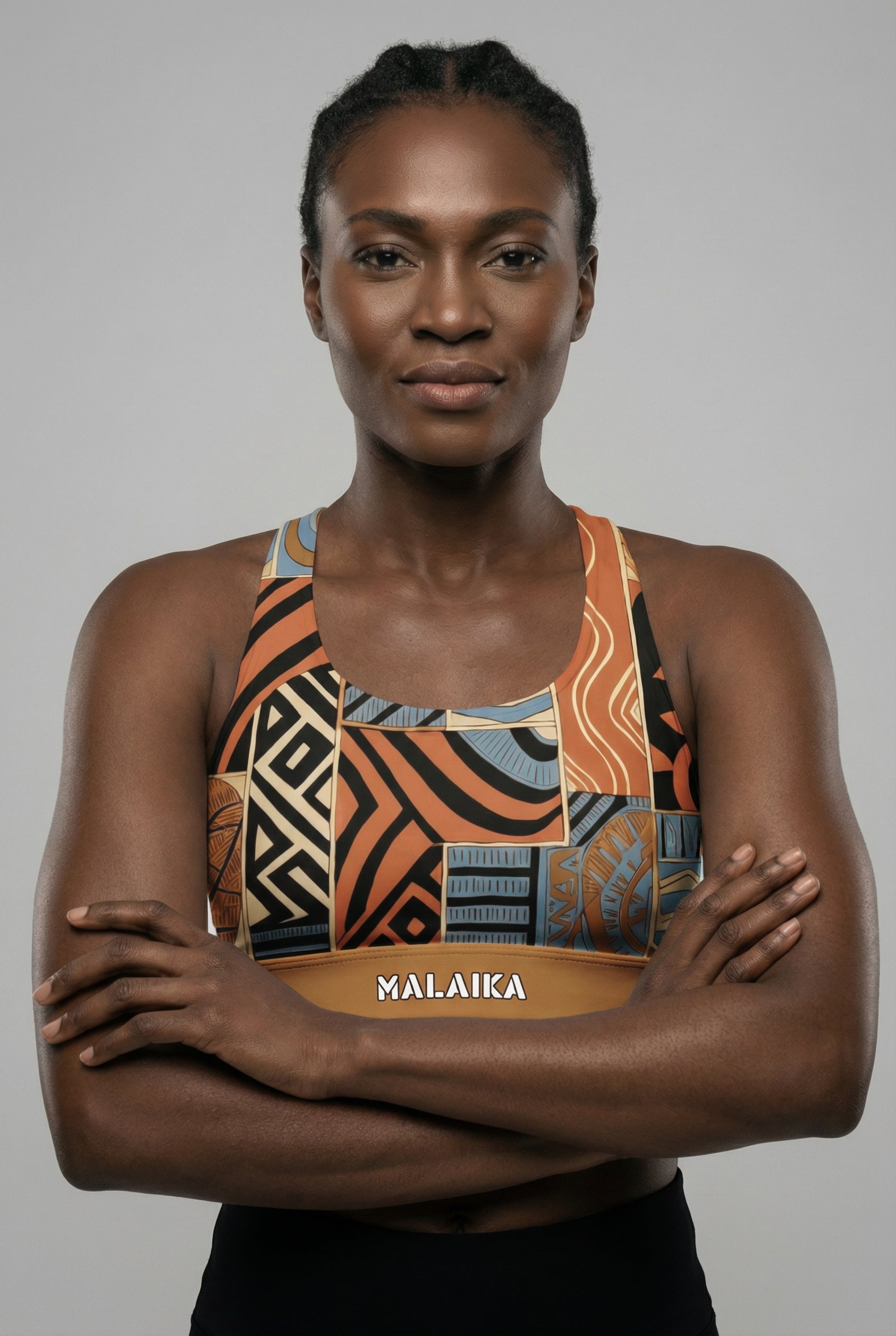 Front view of a fit model flexing biceps, wearing a bold orange and black abstract tribal sports bra with 'MALAIKA' printed on the golden waistband.