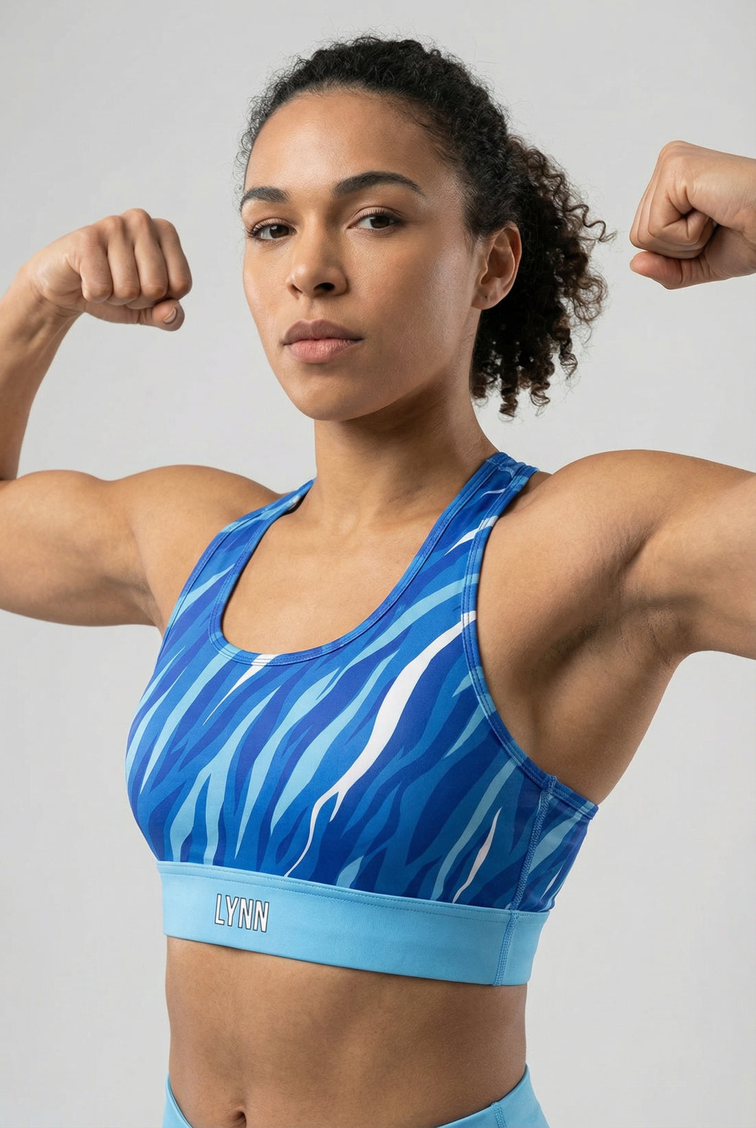 Front view of a sports bra with blue, navy, and white tiger stripes, featuring 'LYNN' printed on the solid sky blue waistband.