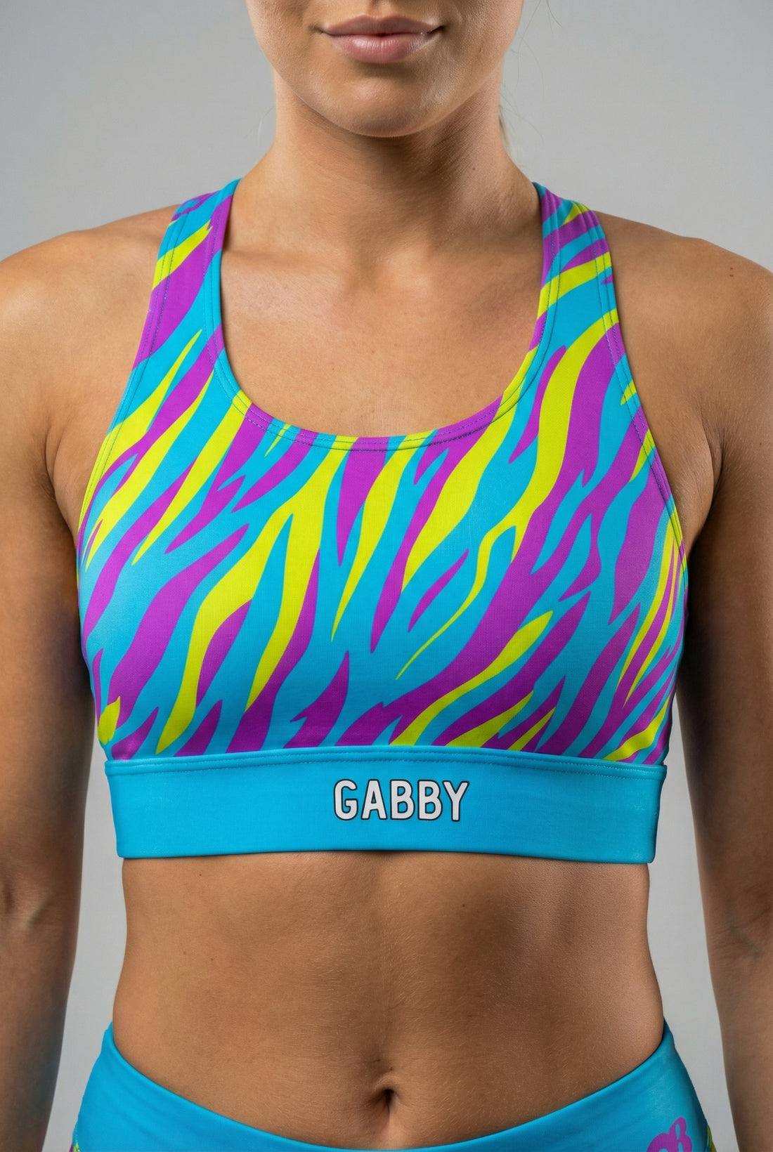 Front view of a confident woman wearing a vibrant cyan, neon yellow, and purple tiger stripe sports bra, featuring 'GABBY' printed on the solid cyan blue waistband.
