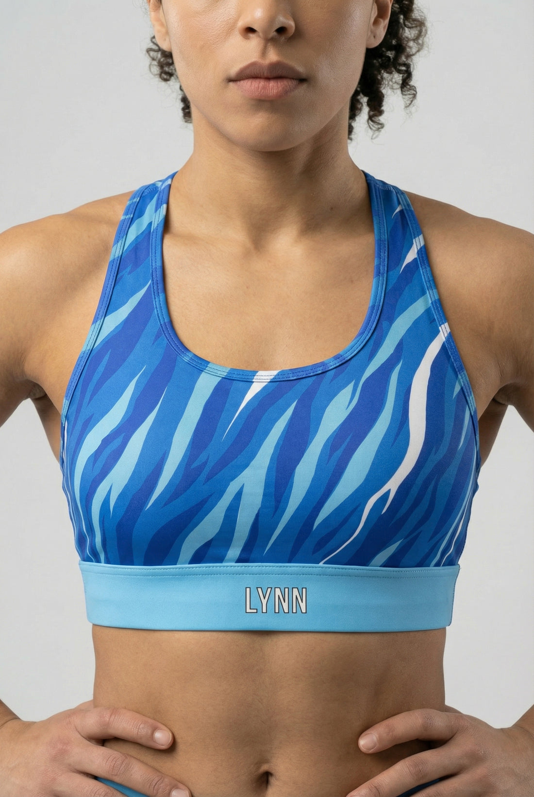 Front view of a sports bra with blue, navy, and white tiger stripes, featuring 'LYNN' printed on the solid sky blue waistband.