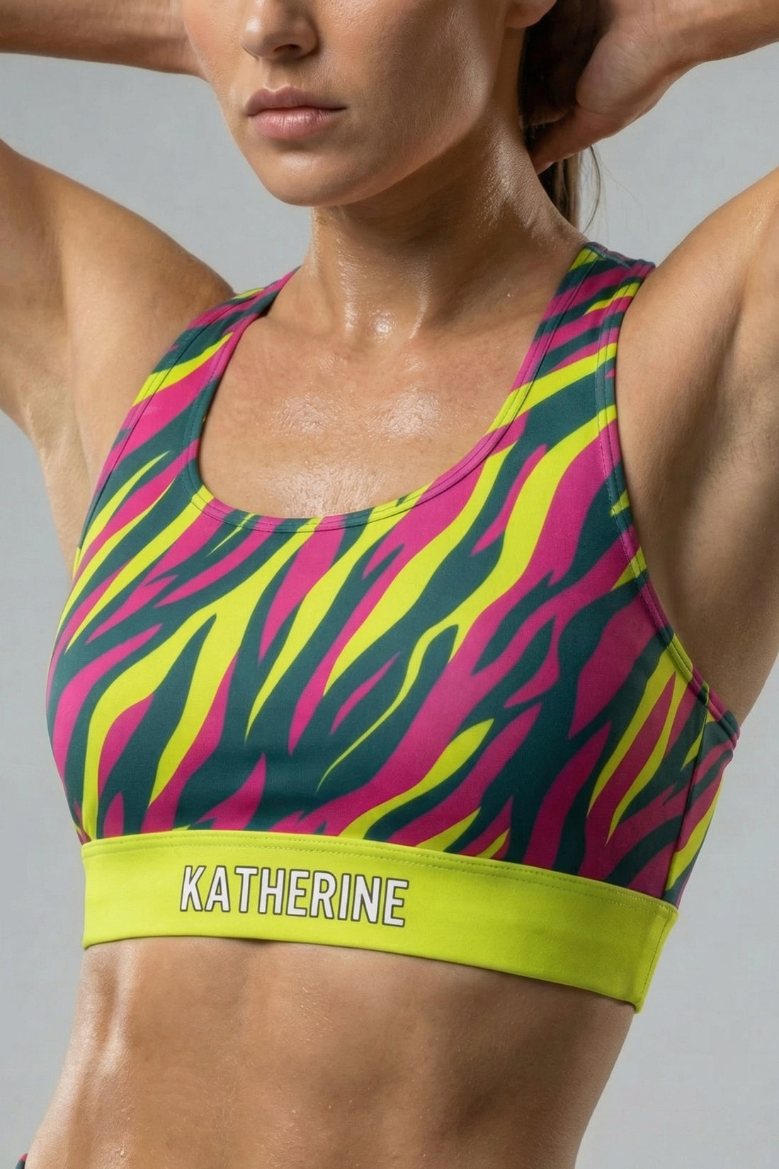 Front view of a confident woman wearing a hot magenta, yellow, and teal tiger stripe sports bra, featuring 'KATHERINE' printed on the solid neon lime waistband.