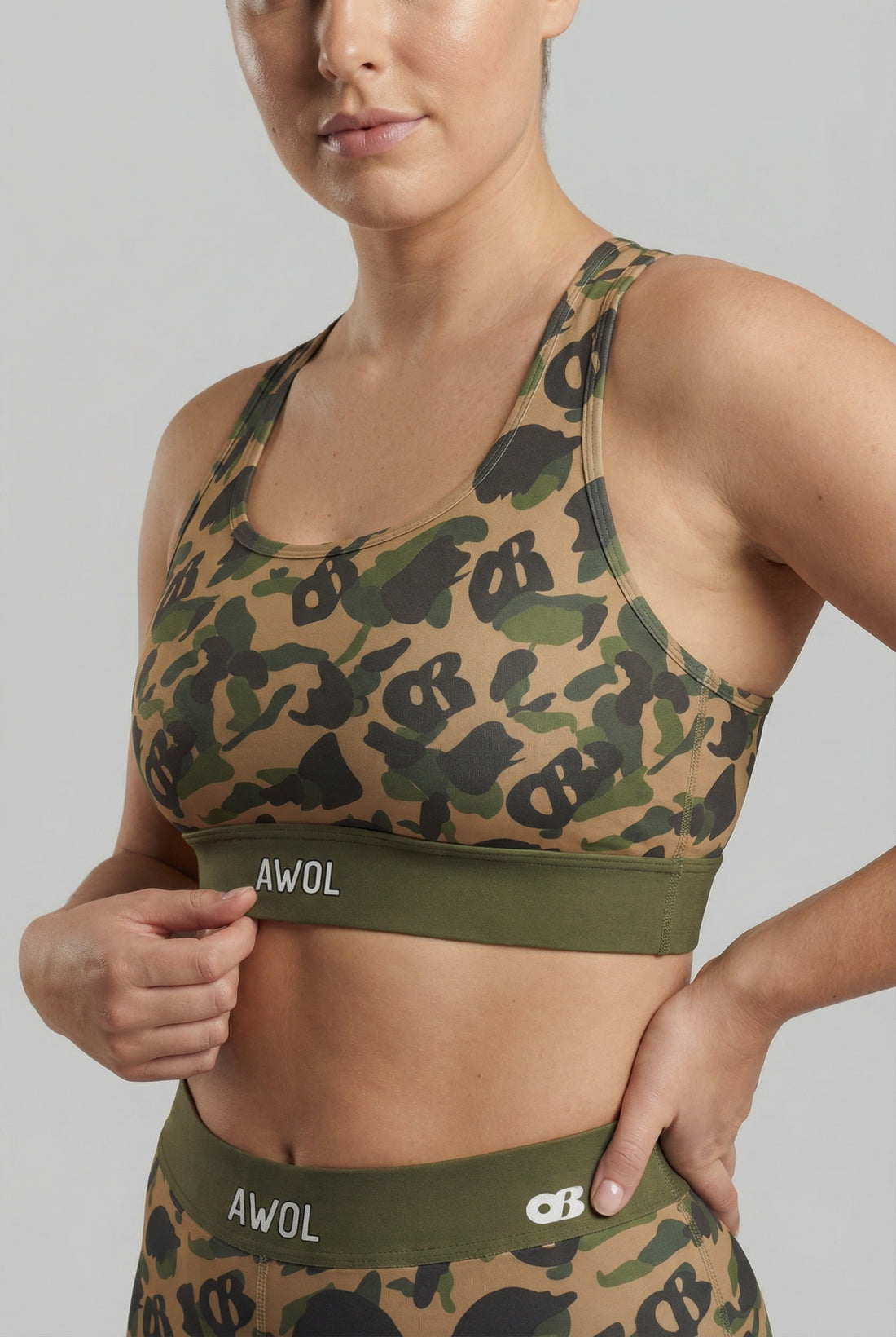 Front view of green camouflage sports bra featuring hidden 'OB' monogram shapes in olive, tan, and black, with 'AWOL' printed on the olive green waistband.