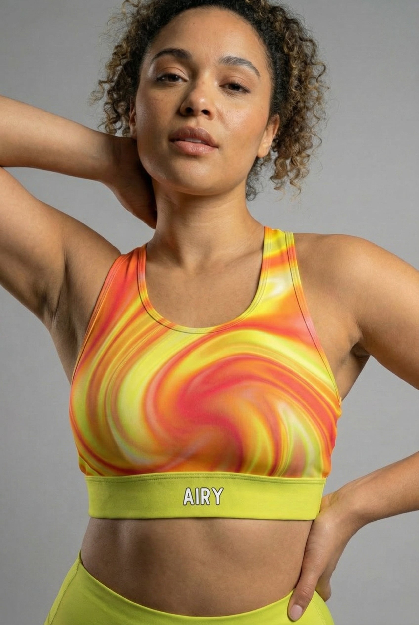 Front view of tie-dye sports bra in orange, yellow, and red swirls, featuring 'AIRY' printed on the neon lime green waistband.