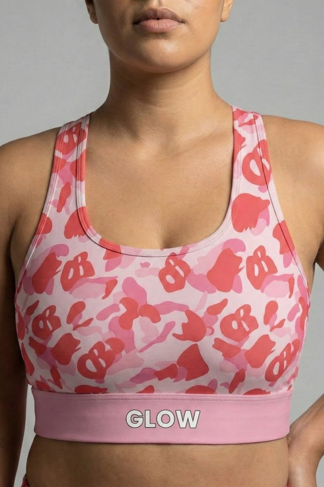 Close-up front view of pink camouflage sports bra featuring hidden 'OB' monogram shapes in red, with 'GLOW' printed on the pastel pink waistband.
