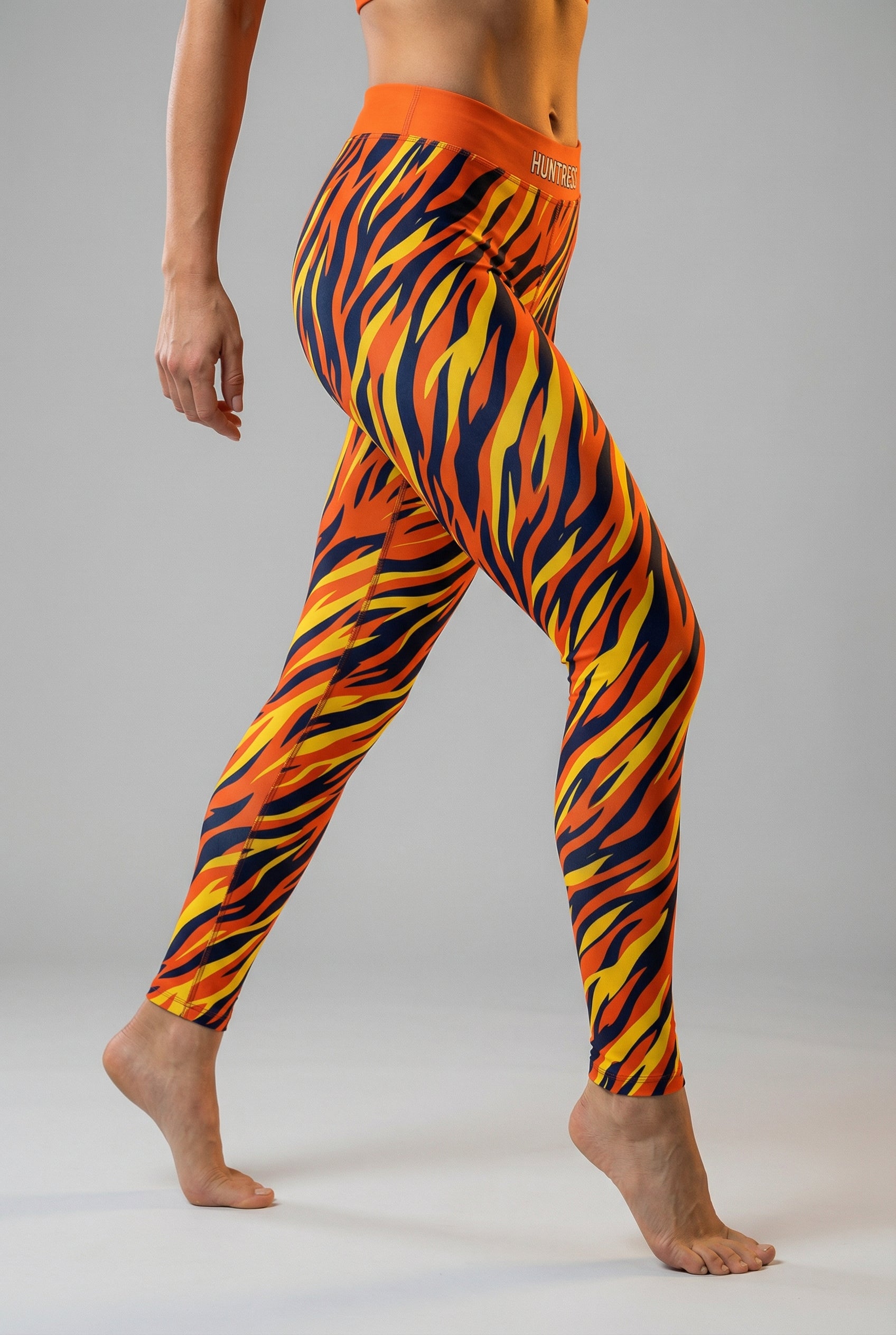 Front view of high-waisted leggings featuring an orange, yellow, and navy tiger stripe pattern, with 'HUNTRESS' printed on the solid orange waistband.