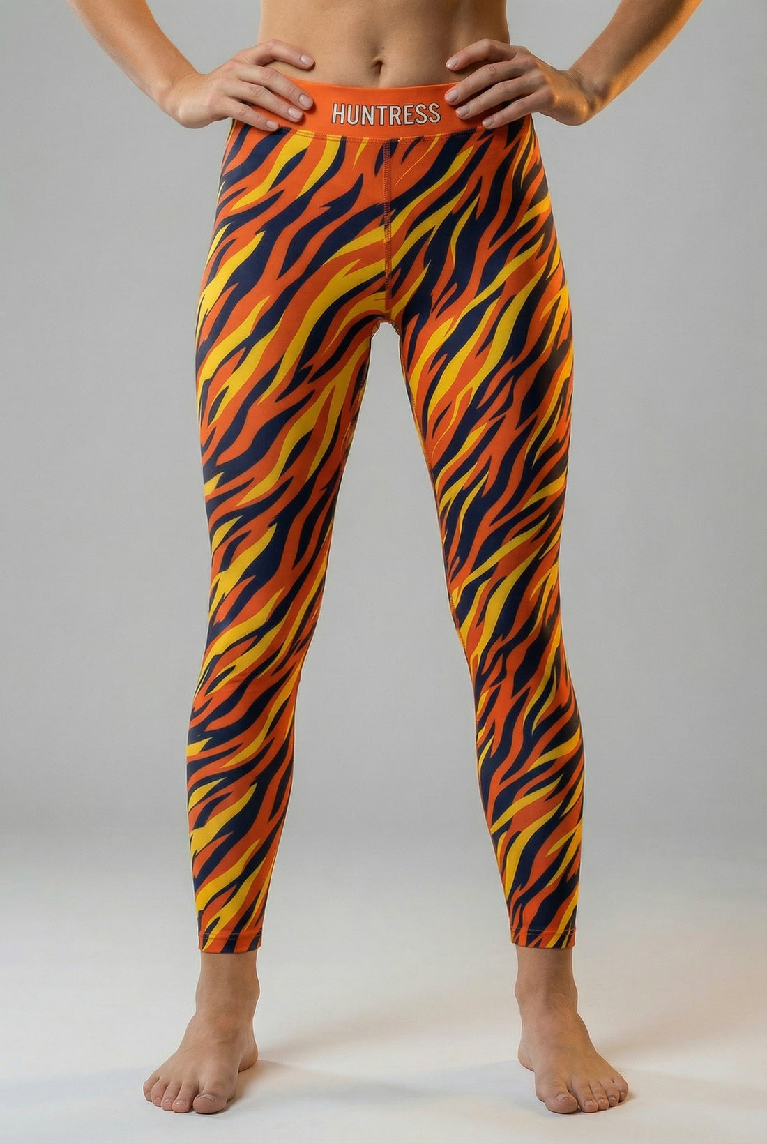 Front view of high-waisted leggings featuring an orange, yellow, and navy tiger stripe pattern, with 'HUNTRESS' printed on the solid orange waistband.