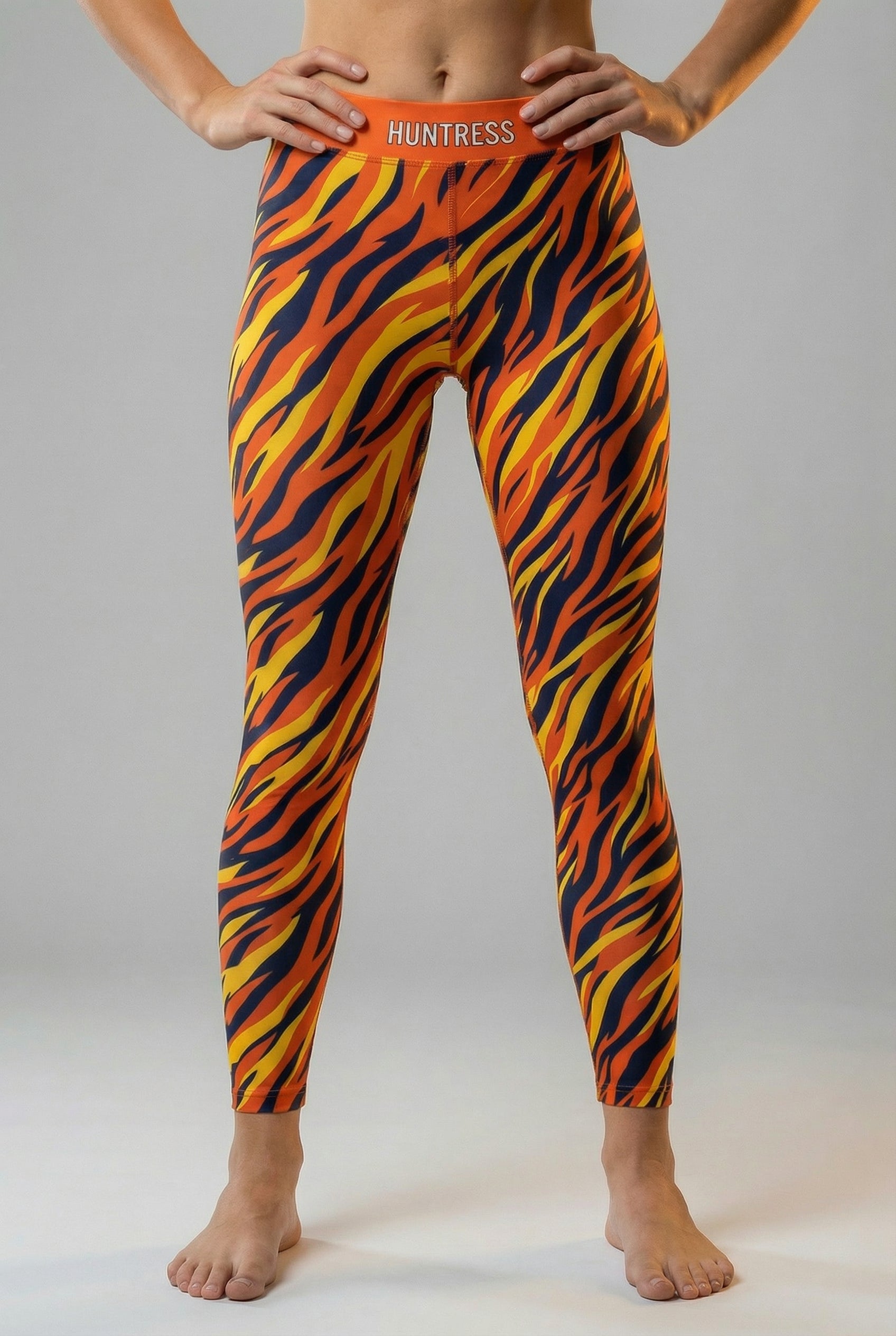 Front view of high-waisted leggings featuring an orange, yellow, and navy tiger stripe pattern, with 'HUNTRESS' printed on the solid orange waistband.
