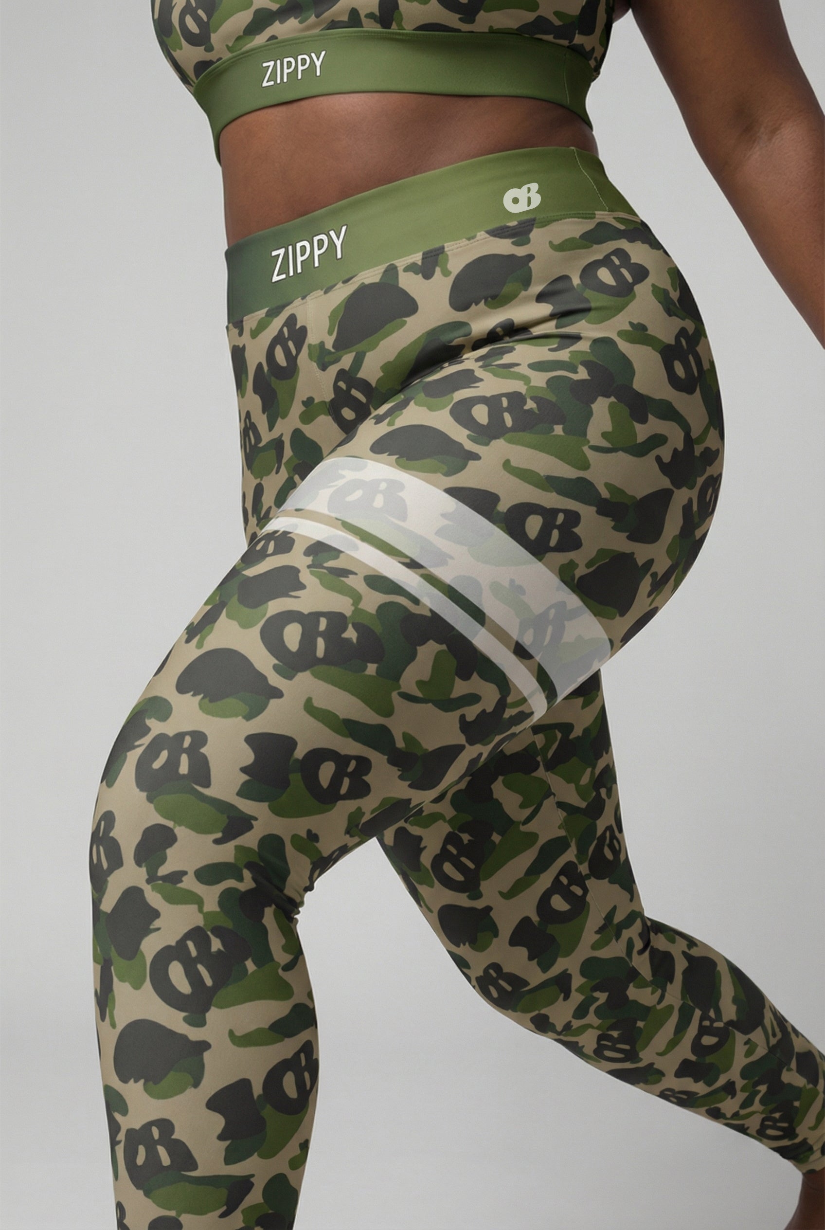 Front view of military green camouflage leggings with hidden 'OB' monogram shapes, featuring white stripes on the left thigh and 'ZIPPY' printed on the olive waistband.