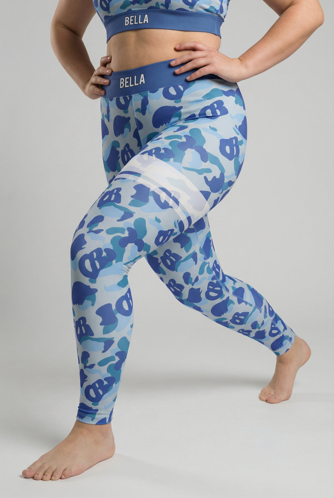 Front view of blue camouflage leggings with hidden 'OB' monogram shapes, featuring white stripes on the left thigh and 'AMA' printed on the slate blue waistband.